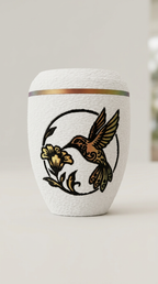 Weiße Urne mit Kolibri-Motiv und Blüte in warmen Gold- und Rottönen, daneben rote Blüten und eine Kerze